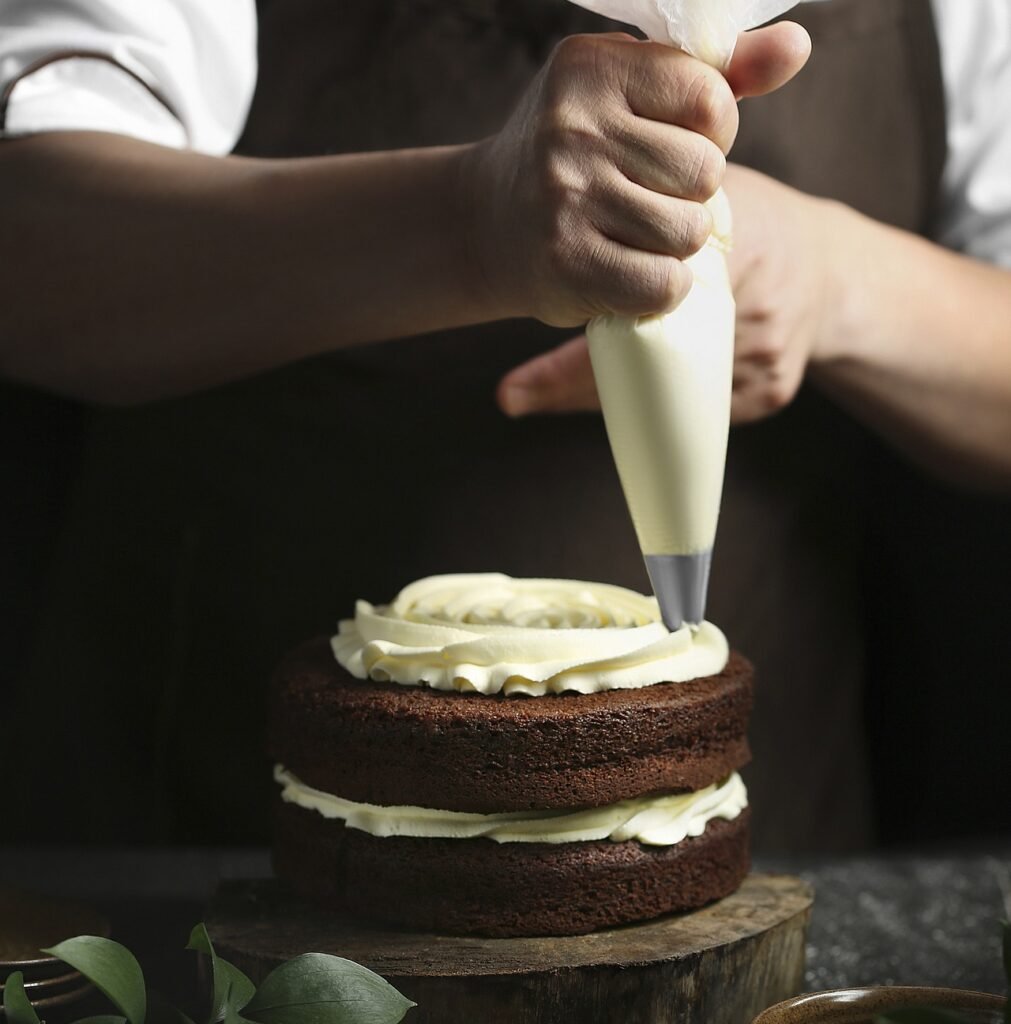 A person carefully decorates a two-layered chocolate cake with cream using a piping bag, adding a personal touch to this homemade chocolate cake.