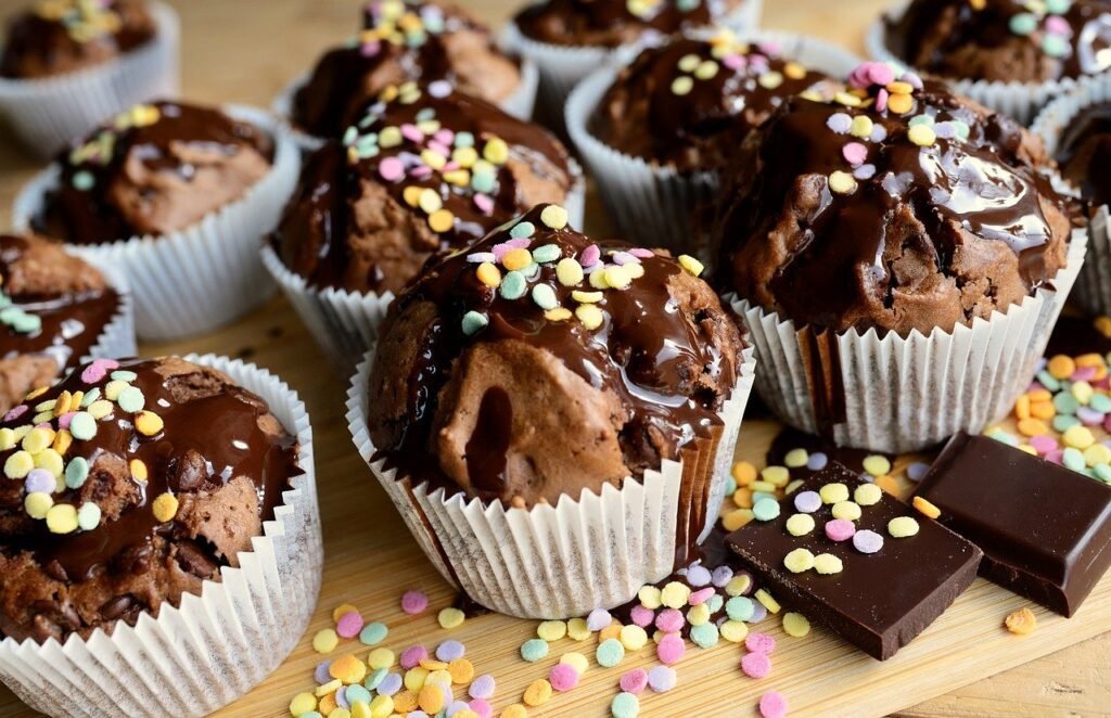 A tempting array of chocolate muffins topped with rich chocolate glaze and vibrant colorful sprinkles, placed on a wooden surface. Accompanied by pieces of dark chocolate, this scene captures the essence of a home baking shop. Perfect for those searching for a cake shop near me, our affordable cakes near me include the best homemade chocolate cake and these delightful homemade chocolate muffins.