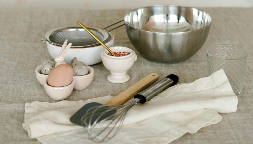 A collection of kitchen utensils and items arranged on a beige cloth, ready for a home baking session. Featuring a mixing bowl, whisk, spatula, and a cute ceramic egg holder, this setup is perfect for those seeking a cake shop near me or affordable cakes near me. Our home baking shop is renowned for the best homemade chocolate cake and a variety of delightful homemade chocolate cakes. Experience the joy of home baking with our quality tools and delicious recipes.
