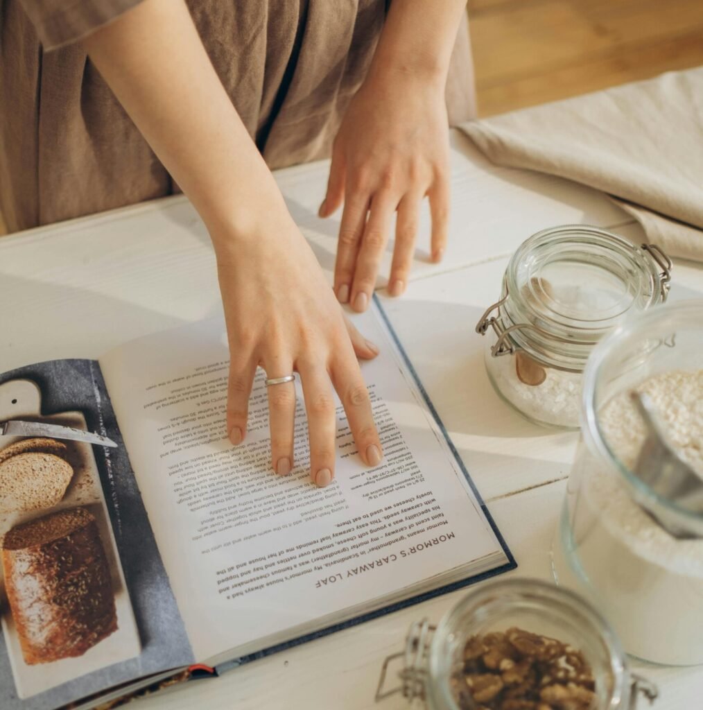 A cookbook open to a recipe with hands preparing ingredients such as flour and nuts. This home baking scene captures the essence of our shop, perfect for those searching for a cake shop near me or affordable cakes near me. Known for the best homemade chocolate cake and other delightful treats, our home baking shop offers a variety of homemade chocolate cakes and baked goods.