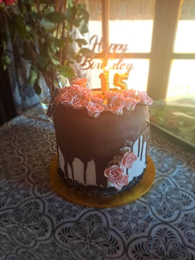 A beautifully decorated birthday cake with a chocolate drip design and pink roses, set against a patterned tablecloth. This home baking masterpiece is perfect for anyone looking for a cake shop near me. Our affordable cakes near me are ideal for all celebrations. Known for the best homemade chocolate cake, our home baking shop offers a delightful array of treats, including this stunning homemade chocolate cake, ideal for special occasions.